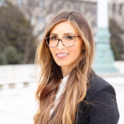 A photo of a white woman with long dark hair and glasses dressed in professional attire